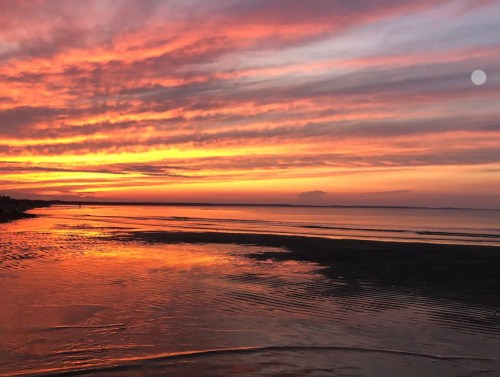 A vibrant sunset over calm waters, with streaks of orange, pink, and purple in the sky and a small moon visible.