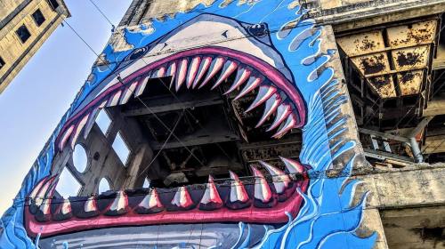 A large mural of a shark's open mouth on a weathered building, with blue waves and exposed structural elements.
