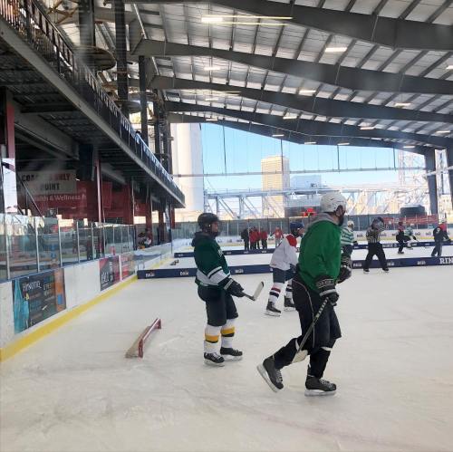 Two hockey players on the ice in a rink, with spectators and a large window showing a cityscape in the background.
