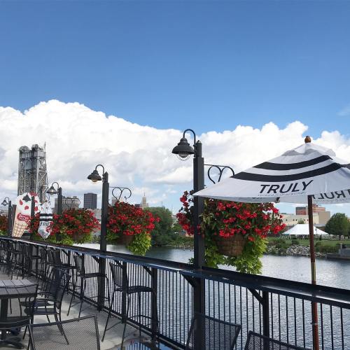 A riverside view with hanging flower baskets, umbrellas, and a bridge in the background under a blue sky with clouds.