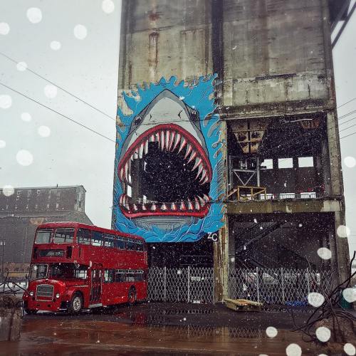 A large shark mural on a concrete building, with a red double-decker bus in front, set in a snowy urban environment.