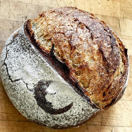 A round loaf of artisan bread with a crusty exterior and a decorative flour pattern on top, resting on a wooden surface.