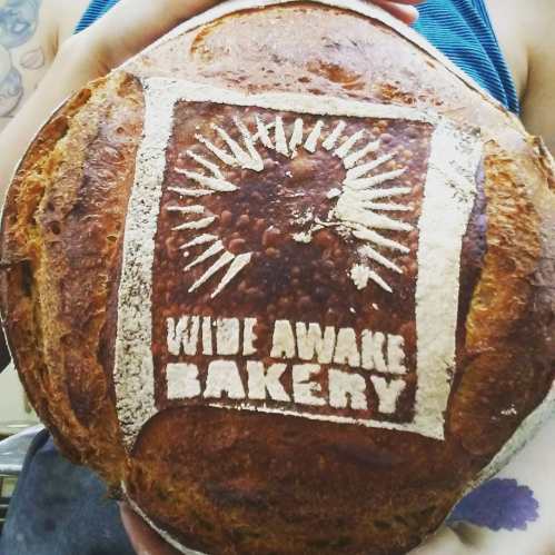 A person holds a round loaf of bread with a logo that reads "Wide Awake Bakery" imprinted on the crust.