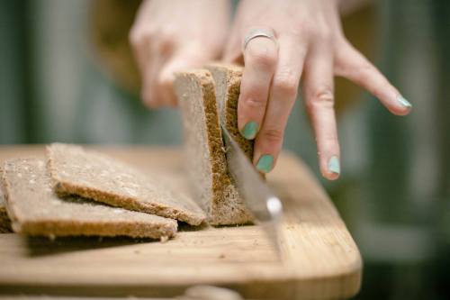 A person with mint green nails slices a loaf of bread on a wooden cutting board.