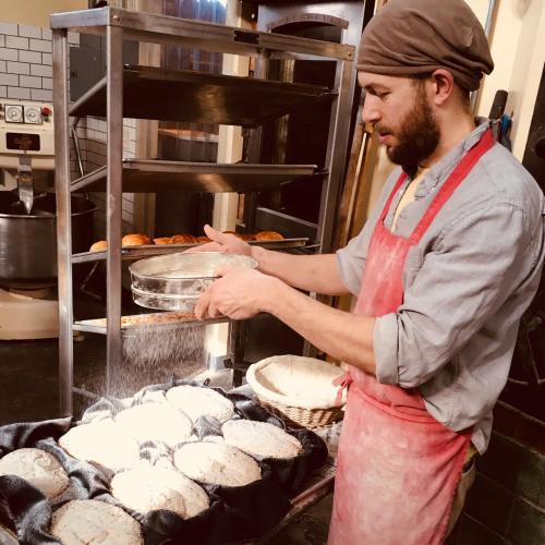 A baker in an apron sifts flour over round loaves of bread on a table, with a rack of baked goods in the background.