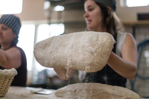 A baker dusts flour over a loaf of bread while another baker works in the background.