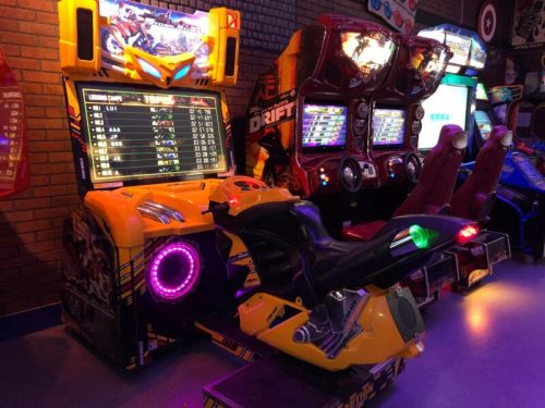 A colorful arcade game setup featuring a motorcycle simulator and racing games against a brick wall backdrop.