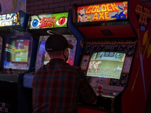 A person playing a retro arcade game at a gaming venue, with "Golden Axe" and "Altered Beast" machines visible.