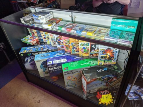 Display case filled with various video games and consoles, including Nintendo 64 and Game Boy, with a sale sign.