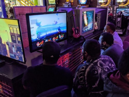 A group of people watching a video game on a large screen in an arcade, with colorful lights and game consoles around.