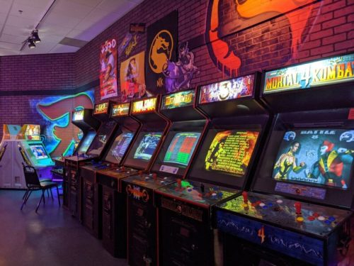 A row of vintage arcade machines in a colorful, dimly lit gaming room with retro posters on the walls.