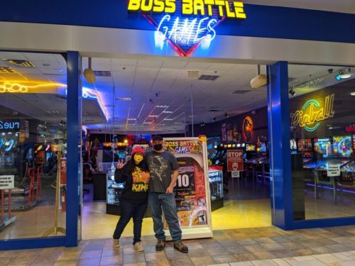 A couple stands outside Boss Battle Games arcade, smiling and posing with arcade machines visible in the background.