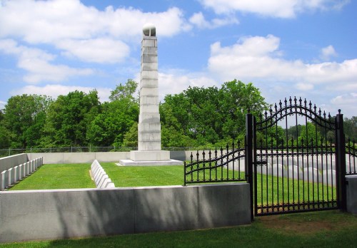 A tall stone monument surrounded by a black wrought-iron fence and green grass, under a partly cloudy sky.