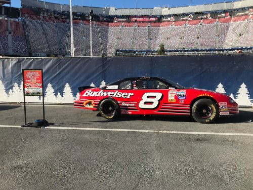 A red NASCAR car with Budweiser branding parked beside a sign in an empty stadium.
