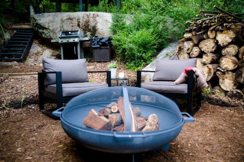 Cozy outdoor seating area with two chairs, a fire pit, and a grill surrounded by greenery and stacked firewood.