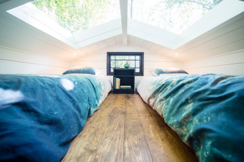 Cozy bedroom with two beds, large skylights, and a small bookshelf, surrounded by greenery.