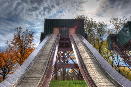 A large wooden slide structure with two steep slides, surrounded by trees and a cloudy sky.