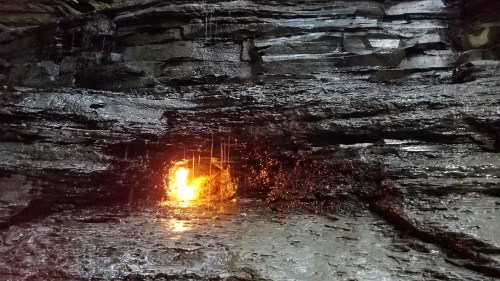 A glowing orange light emerges from a dark, rocky crevice, with water droplets cascading down the wet surface.