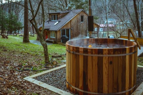 A wooden hot tub sits outdoors near a cozy cabin, surrounded by trees and a grassy area. Steam rises from the tub.