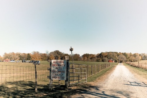 A gravel path leads to a field with a sign prohibiting alcohol, profanity, pets, and video cameras. Trees in the background.