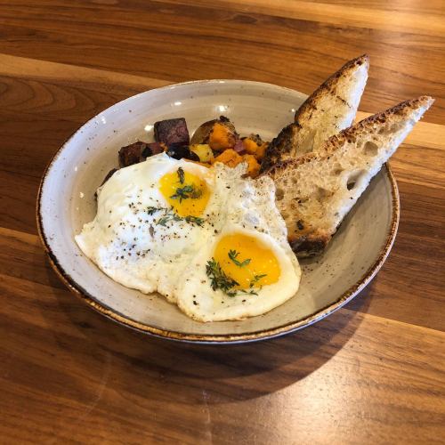A bowl of two sunny-side-up eggs, roasted potatoes, and two slices of toasted bread on a wooden table.