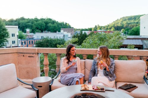 Two women sit on a rooftop terrace, enjoying drinks and chatting, with a scenic view of a town and greenery in the background.