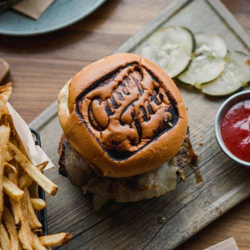A close-up of a burger with a branded bun, served with fries, pickles, and a small bowl of ketchup on a wooden board.