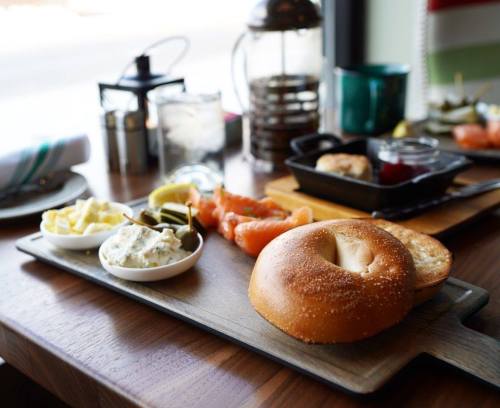 A wooden platter with a bagel, cream cheese, smoked salmon, pickles, and a side of assorted condiments.