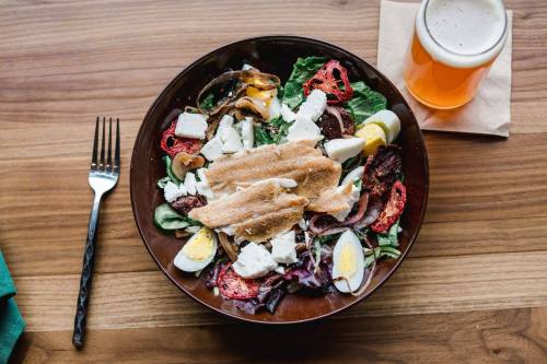 A colorful salad with greens, grilled vegetables, eggs, and feta cheese, alongside a glass of beer on a wooden table.