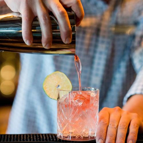 A bartender pours a pink cocktail from a shaker into a glass, garnished with a lime slice.