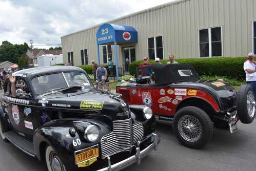 Two classic cars parked in front of a building, with spectators nearby enjoying a car show event.