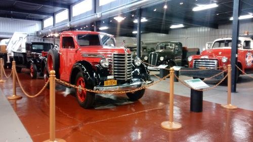 A vintage red truck on display in a museum, surrounded by other classic vehicles and a polished floor.