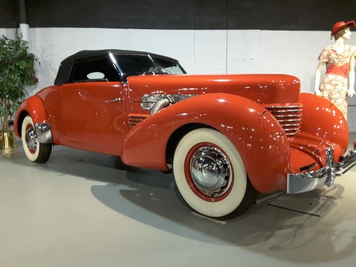 A vintage red convertible car with a black top, displayed in a museum setting alongside a mannequin in a dress.
