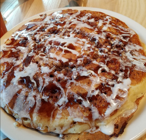 A large, fluffy cinnamon roll pancake drizzled with icing on a white plate.