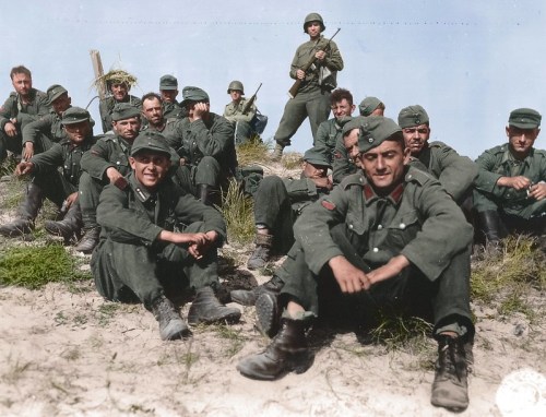A group of soldiers in green uniforms, some sitting and others standing, on a grassy terrain, in a historical colorized photo.