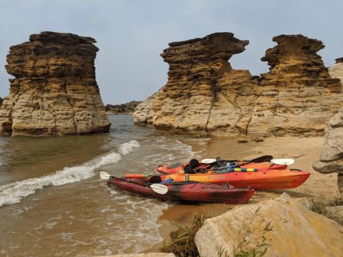 Two kayaks on a sandy beach near rocky cliffs, with calm water and a hazy sky in the background.