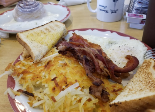 A plate of breakfast featuring eggs, crispy bacon, hash browns, and toast, with a cinnamon roll in the background.