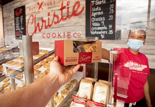 A hand holds a box of cookies in front of a cookie shop, with a smiling employee in a mask behind the counter.
