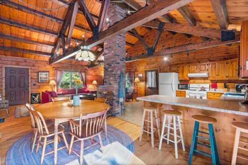 Cozy cabin interior with wooden beams, a round dining table, and a living area featuring a fireplace and kitchen.