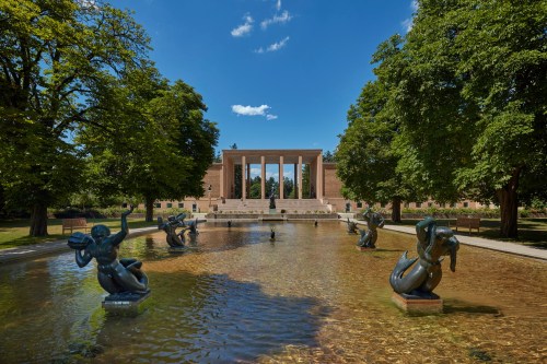 A serene park scene featuring a pond with sculptures, surrounded by trees and a grand building in the background.