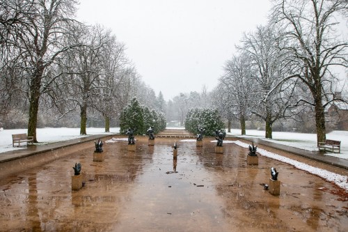 A snowy park scene featuring a frozen pond, bare trees, and sculptures, creating a serene winter landscape.