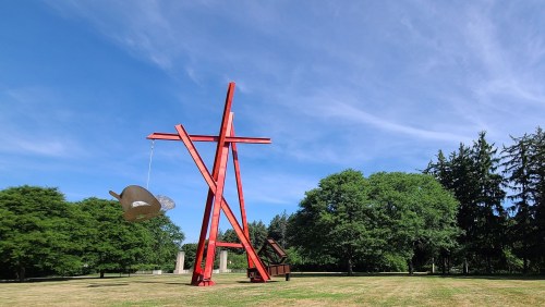 A large red sculpture resembling a balance scale stands in a grassy park surrounded by trees under a blue sky.