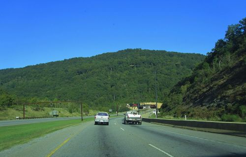Two trucks driving on a highway with green hills in the background under a clear blue sky.