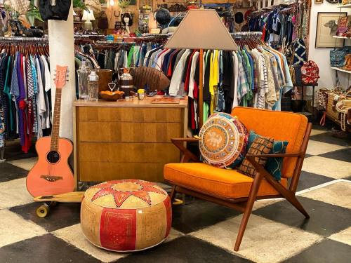 A cozy corner with an orange chair, patterned pouf, guitar, and a lamp, surrounded by colorful clothing racks.