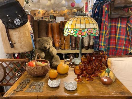 A cozy vintage shop display featuring a teddy bear, a stained glass lamp, fruit, and amber glassware on a wooden table.