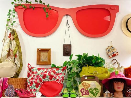 Colorful display of vintage accessories, including bags and hats, with large red sunglasses on the wall above.
