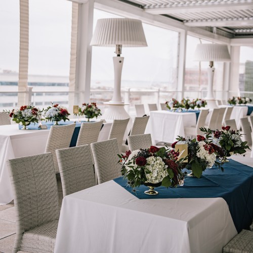 Elegant dining setup with white and blue tablecloths, floral centerpieces, and large lamps in a bright, airy space.