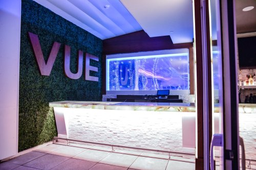 Modern bar area with a green wall, illuminated counter, and a blue-lit backdrop featuring flowing water.