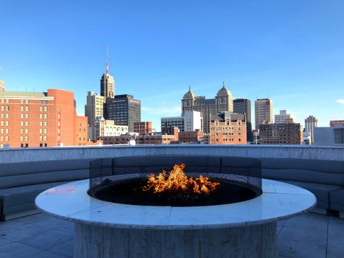 A rooftop view featuring a fire pit, with city skyscrapers and a clear blue sky in the background.
