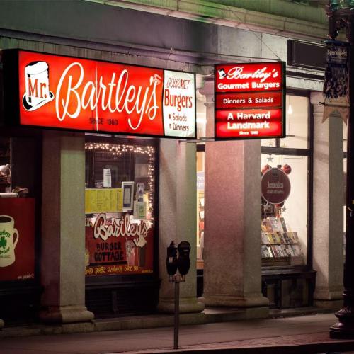 Brightly lit restaurant signs for Mr. Bartley's Gourmet Burgers, showcasing burgers, salads, and a Harvard landmark.